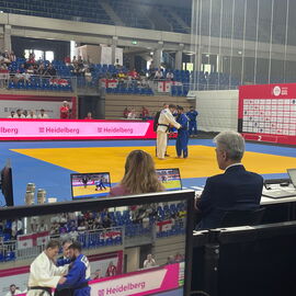 Blick über die Schulter der Juror*innen auf Bildschirme bei der Para-Judo-Europameisterschaft.  Zwei Judoka kämpfen.