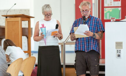 In der Aula stehen eine Frau und ein Mann mit Mikrofonen in der Hand, sie hält eine Schultüte.