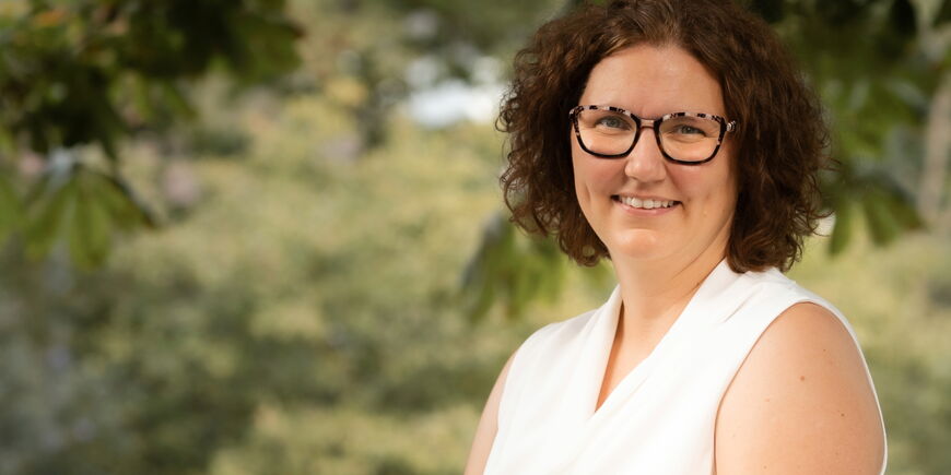 Portraitfoto  Melanie Grzimbke - sie hat braune Locken und trägt eine Brille. Sie trägt ein weißes Oberteil.