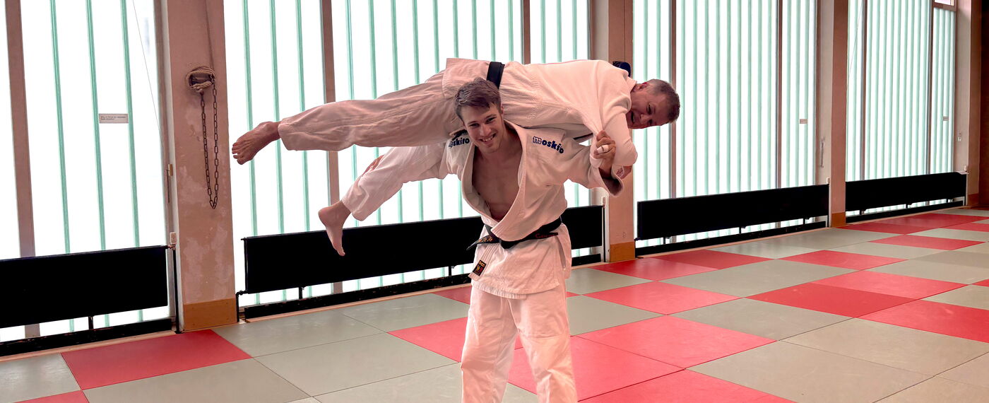 Zwei Judotrainer, die eine dynamische Judotechnik in einer Turnhalle auf Matten vorstellen.