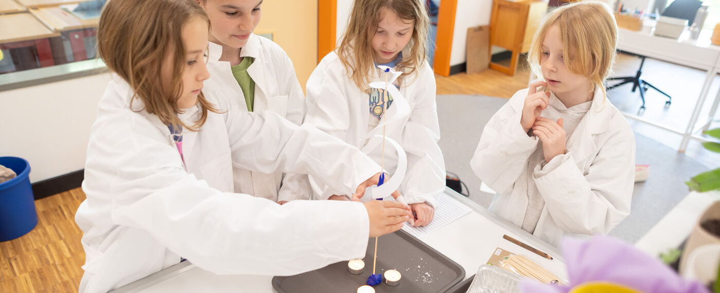 Vier Schüler*innen in weißen Kitteln stehen an einem Labortisch und halten eine Papierspirale über Teelichter.