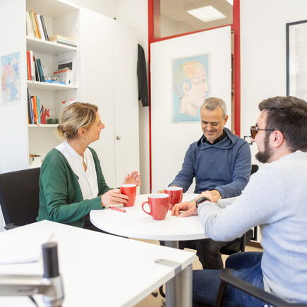 Drei Menschen sitzen in einem Büroraum um einen runden Tisch mit Kaffeetassen.