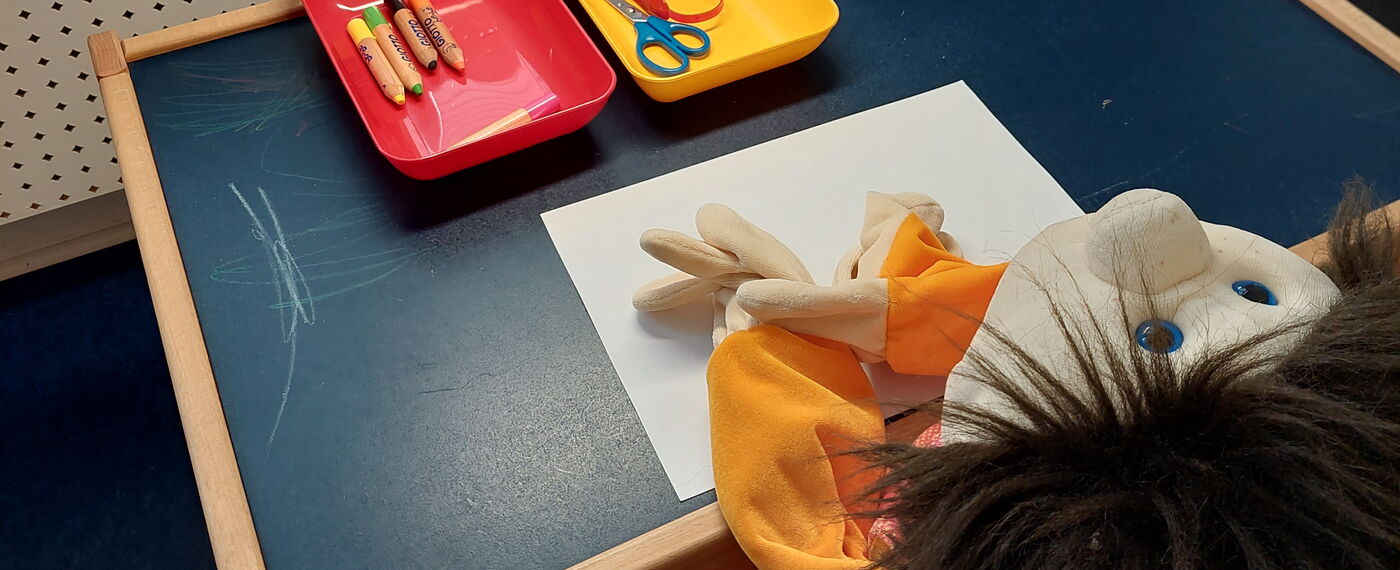 An einem Spieltisch sitzt eine Handpuppe auf einem Kinderstuhl, vor sich ein weißes Blatt Papier und Stifte und Schere in bunten Aufbewahrungsschalen.