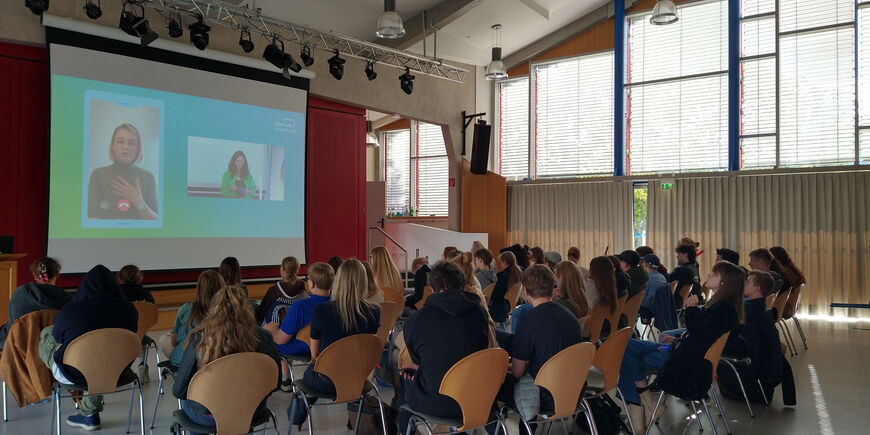 In der abgedunkelten Aula sitzen Schüler*innen auf Stühlen vor einer großen Leinwand und folgen einer Online-Veranstaltung.