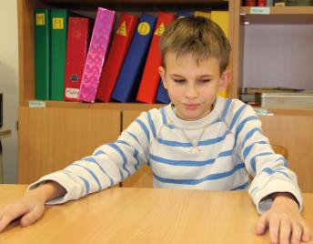 Das Foto zeigt einen Jungen, der - seine Arme breit aufgestützt - lächelnd am Schreibtisch sitzt.