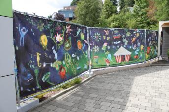 Den rund 15 Meter langen Bauzaun für die Erweiterung der Montessori-Schule auf dem Gelände der Deutschen Blindenstudienanstalt ziert die Reproduktion des Gemäldes „Sekundenschlaf“ der Marburger Künstlerin Uli Krappen. Ein farbenfrohe Traummotiv 