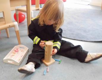 Ein Mädchen aus dem Montessori-Kinderhaus sitzt in Feuerwehr-Uniform vor einem Holzspielzeug.