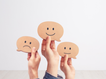 Drei Hände halten Sprechblasen in die Höhe, die mal lächelnde, mal traurige Gesichter zeigen.