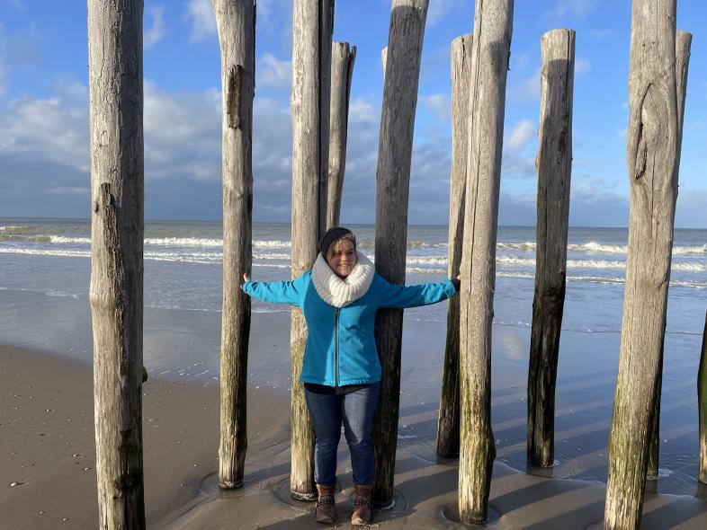 Lea steht an einem irischen Sandstrand zwischen hohen Holzpfählen, die Arme ausgestreckt zu den Seiten, hinter ihr Meer mit Wellen und teils bewölkter blauer Himmel.