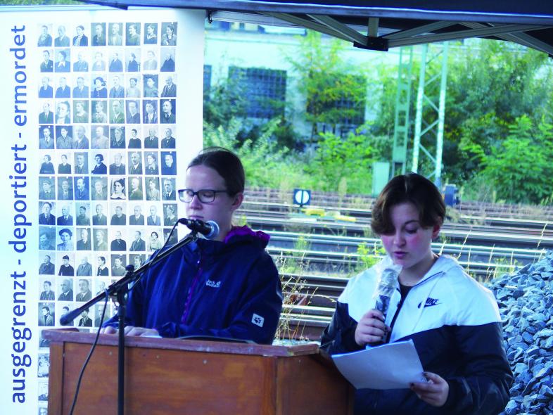 Vanessa und Celina verlesen am Rednerpult die 43 Namen. Im Hintergrund steht links ein Aufsteller mit zahlreichen Schwarzweiß-Porträts von deportierten Menschen, rechts sind die Schienenstränge des Marburger Bahnhofes zu sehen.