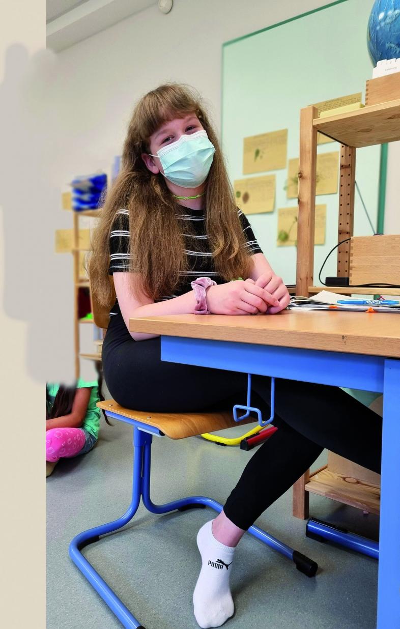 Foto von Madita aus der Lerngruppe 2 der Montessori-Grundschule an einem Tisch im Klassenraum sitzend mit medizinischer Maske.
