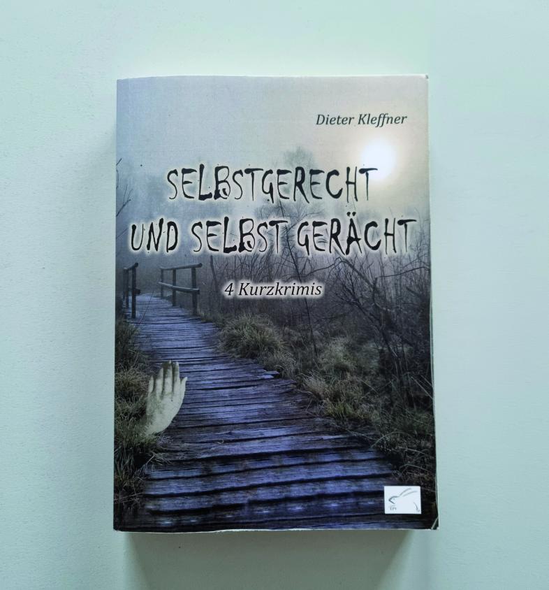 Das Buchcover von "Selbstgerecht und selbst gerächt" zeigt eine weichgezeichnete neblige Moorlandschaft. Eine einzelne Hand ragt neben einem Bohlenweg aus dem Moor, das Licht ist trübe.