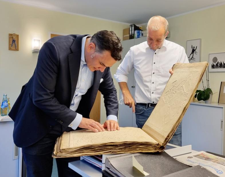 Dr. Stefan Naas befühlt eine historische Karte, die in einem großformatigen Band eingebunden ist. Patrick Temmesfeld steht daneben und hält den Deckel des Buches.