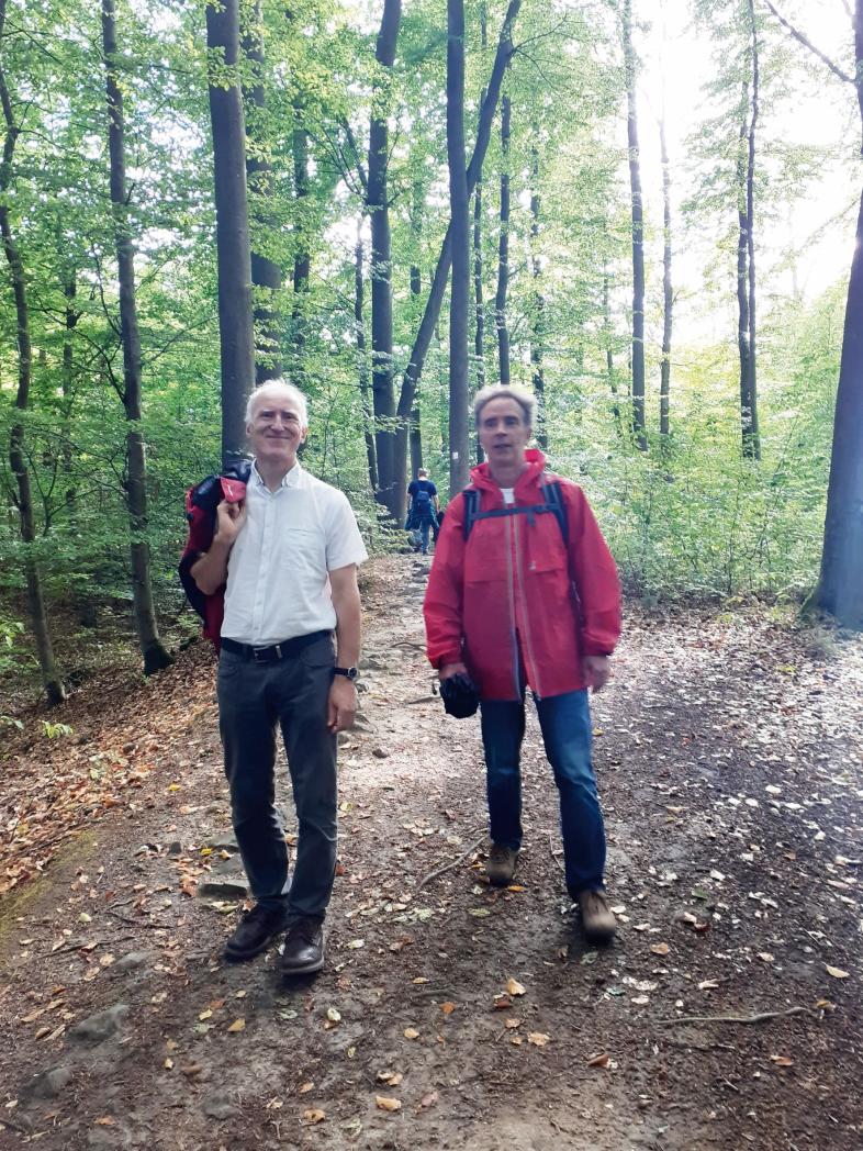 Zwei Lehrer der Carl-Strehl-Schule laufen auf einem breiten Weg durch den grünen Buchenwald. Einer hat die Jacke ausgezogen und über die Schulter gehängt, der Andere trägt eine rote Regenjacke.