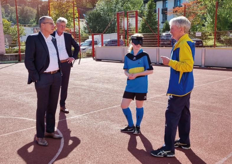 Peter Beuth und Maarten Kubeja stehen auf dem Sportplatz und sprechen mit Manfred Duensing und einer Schülerin in Fußballkleidung und mit Dunkelbrille.