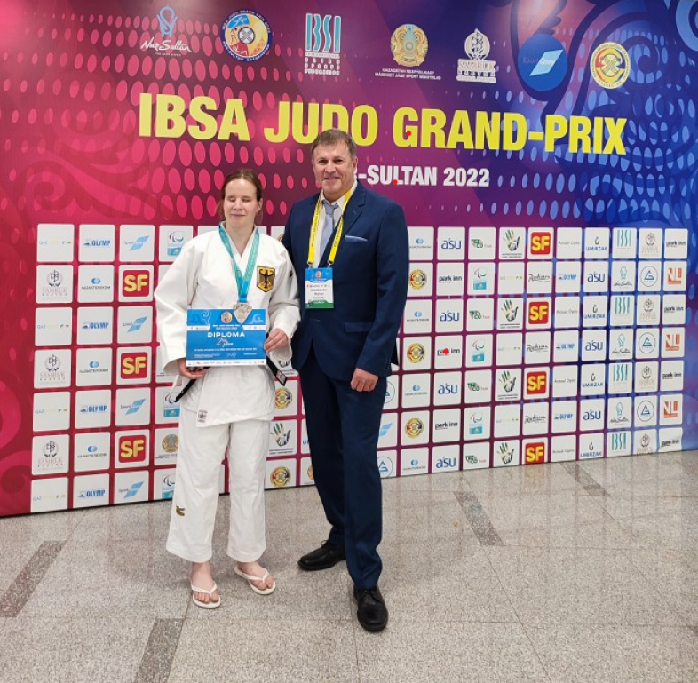 Judoka Vanessa Wagner mit Silbermedaille und Urkunde und Trainer Martin Zaumbrecher posieren strahlend vor einer großen Wand mit dem Judo Grand-Prix Nur-Sultan 2022-Plakat.