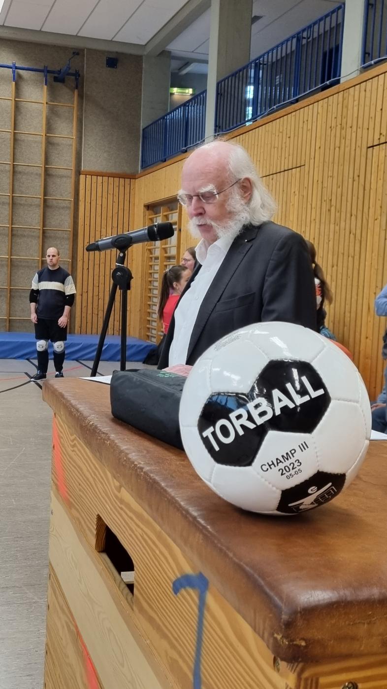 Jürgen Hertlein steht in der blista-Sporthalle vor einem Mikrofon, vor ihm liegt ein Torball.