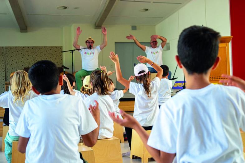 In der Aula zeigen zwei Erwachsene die Choreographie zur Musik, die Kinder tanzen mit.