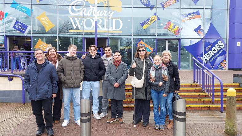 Die Gruppe Schüler*innen und Lehrer*innen steht vor dem Eingang von Cadbury World. Im Hintergrund sind große Werbegrafiken für Süßigkeiten an der Glasfassade zu sehen.
