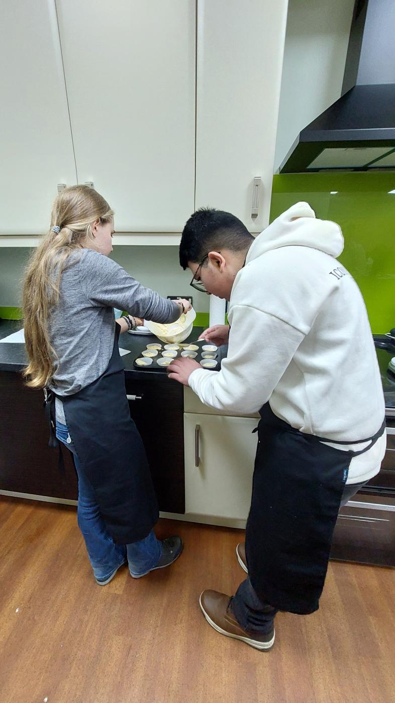 Zwei Personen mit Schürzen füllen in einer Küche Teig in Muffinförmchen. Die Förmchen stehen auf einem Backblech auf der Arbeitsfläche.