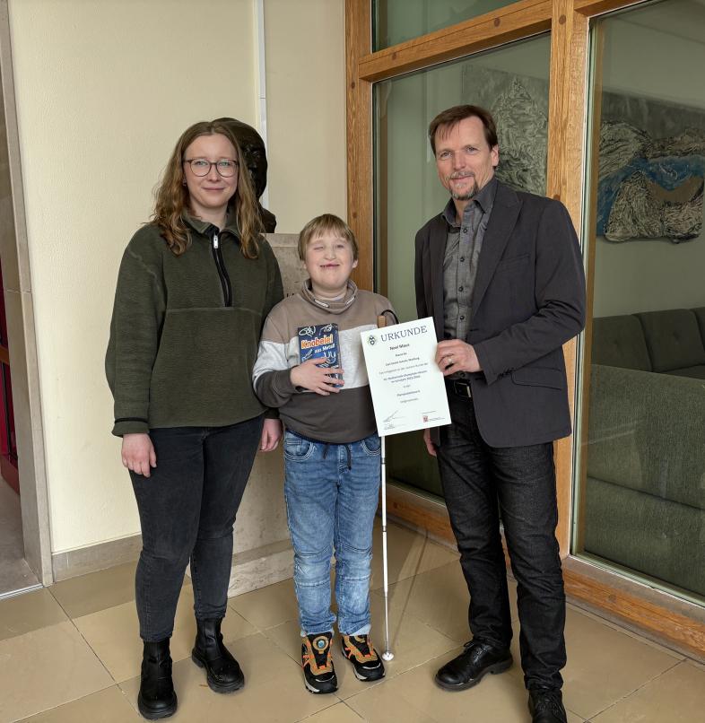 Im Foyer der CSS überreicht Schulleiter Peter Audretsch Noel eine Urkunde gemeinsam mit Debora Debes.