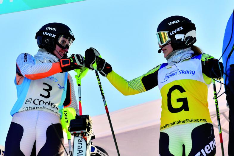 Noemi Ristau und Paula Brenzel in ihrer Rennausrüstung mit Helmen, Skibrillen. Ristau trägt die Startnummer 3, Guidin Brenzel hat eine breite gelbe Markierung mit einem großen schwarzen G um die Körpermitte.