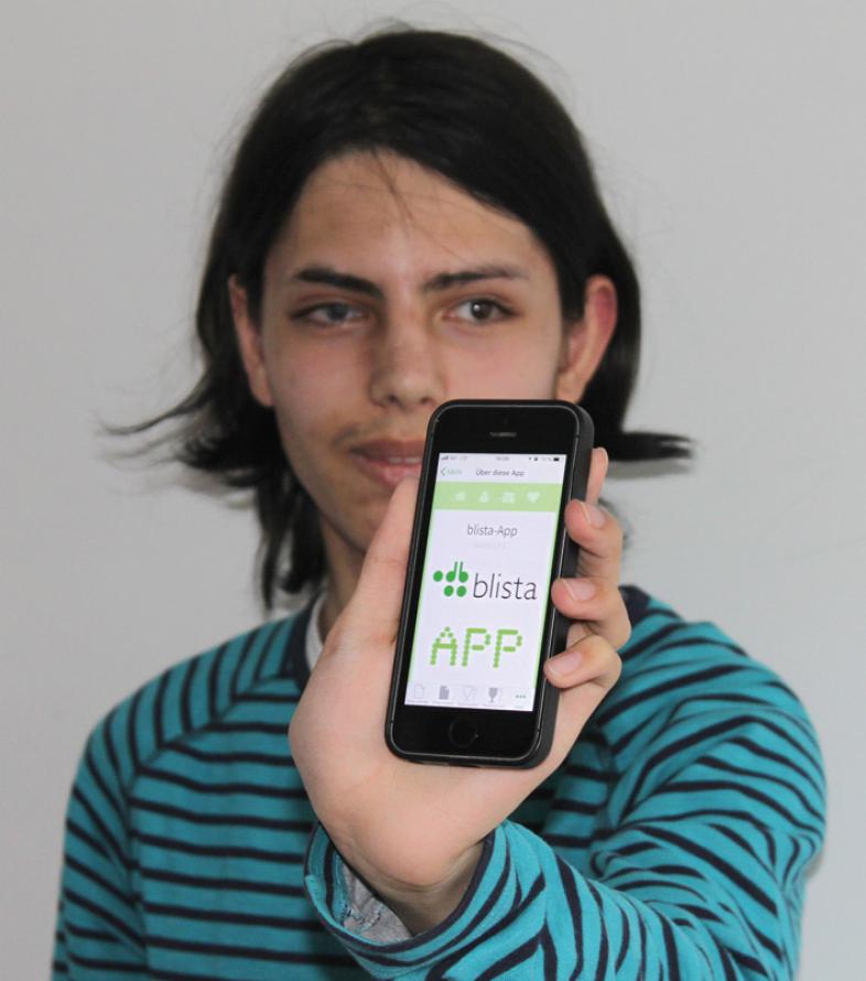 Das Foto zeigt einen jungen Mann mit dunklem mittellangem Haar, er zeigt sein iPhone mit der Anwendung