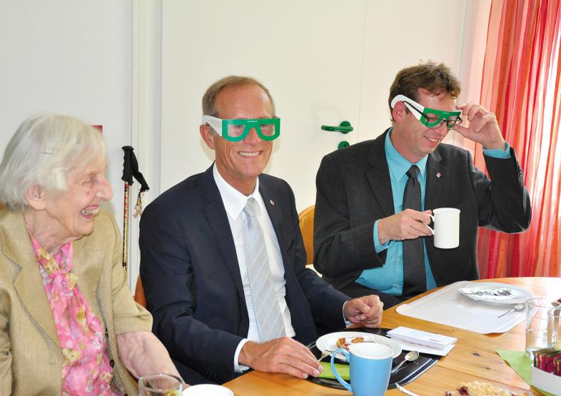 Frau Pfeifer, Staatsminister Wintermeyer und der Regionalbeauftragte für Mittelhessen Scherf (v.l.n.r.). Wintermeyer und Scherf tragen am Kaffeetisch Simulationsbrillen.