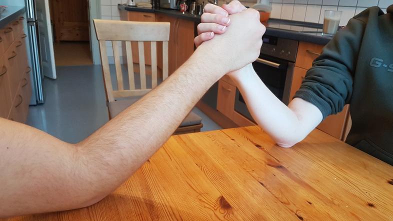 Zwei Arme sind zum Armdrücken gekreuzt auf einen Holztisch abgestützt, im Hintergrund ist eine Küche.
