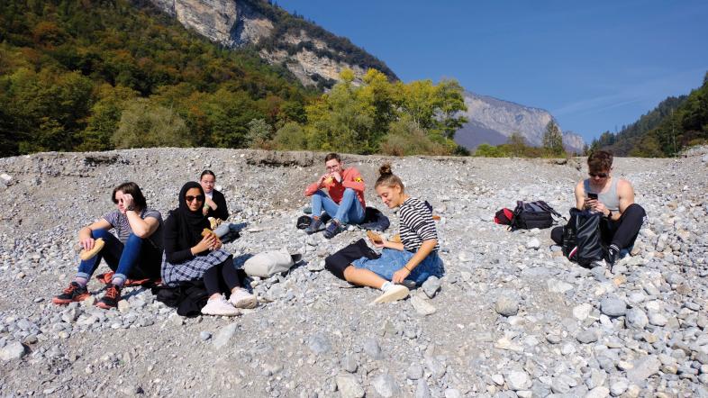 Bild 2: Mittagessen im Flussbett. 6 Schülerinnen und Schüler sitzen im Sonnenschein auf einer riesigen Kiesbank im Flussbett. Im Hintergung sind bewaldtete Hänge und hohe Berge mit steilen Felswänden zu erkennen.