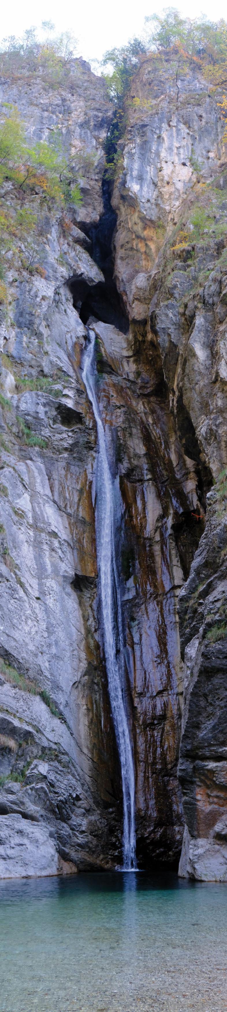 Ein sehr hoher und schmaler Wasserfall ergießt sich zwischen steilen Felswänden in ein glasklares natürliches Wasserbecken mit Kieselgrund.