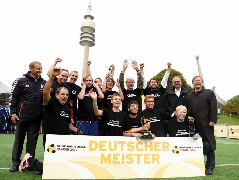 Die Blindenfußballmannschaft jubelt hinter dem "Deutscher Meister"-Schild. Im Hintergrund ragt ein Fernsehturm in den Himmel.