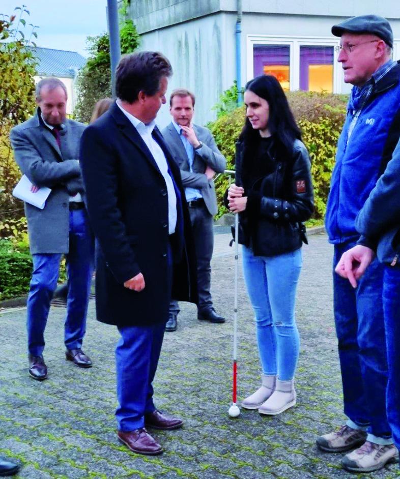 Jürgen Dusel im Gespräch mit einer Schülerin mit Langstock auf einem Weg auf dem blistaCampus. Im Hintergrund stehen Patrick Temmesfeld und Peter Audretsch.