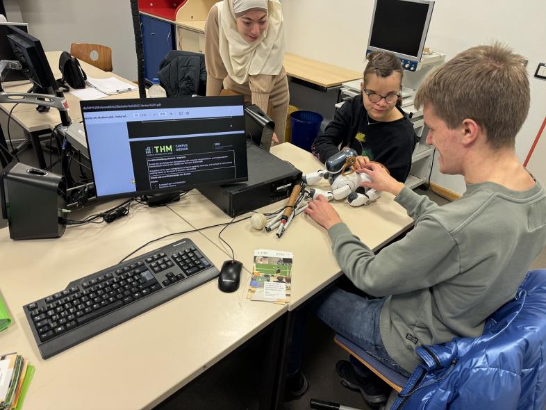 Die drei Schüler*innen Noor, Nejla und Niklas begutachten einen ca. 20 cm großen Roboterhund, der auf einem Tisch in einem Klassenraum liegt. Auf einem der Computerbildschirme ist das Logo der THM zu erkennen. 