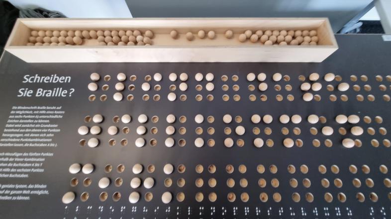 Brailletafel mit Punktschrift-Zeichen. Mittels Holzkugeln, welche in Vertiefungen gesteckt werden, entstehen die gewünschten Punktschrift-Zeichen.