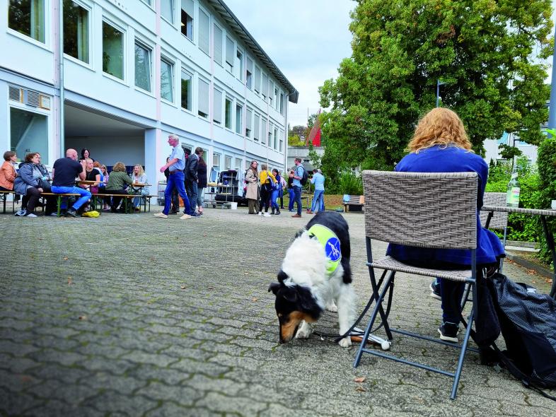 Foto einer Szene vom CampusTag auf dem Schulhof. Im Vordergrund sitzt eine Internatsmitarbeiterin mit Blindenhund an einem Tisch, im Hof laufen einige blista-Mitarbeiter und -Mitarbeiterinnen herum.
