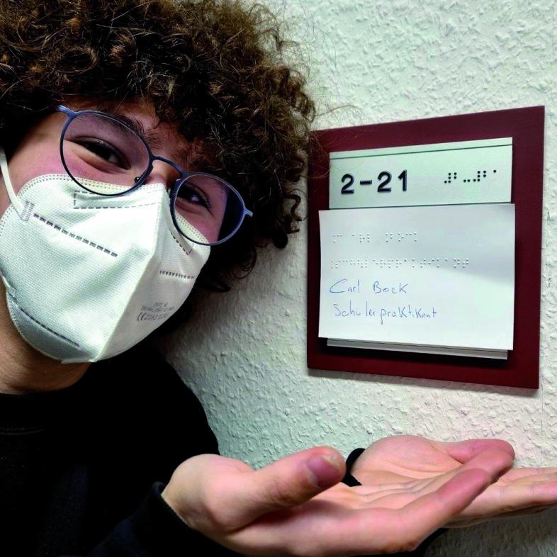 Carl mit dunklen Wuschellocken, Brille und FFP2-Maske präsentiert sein provisorisches, handschriftliches Büroschild
