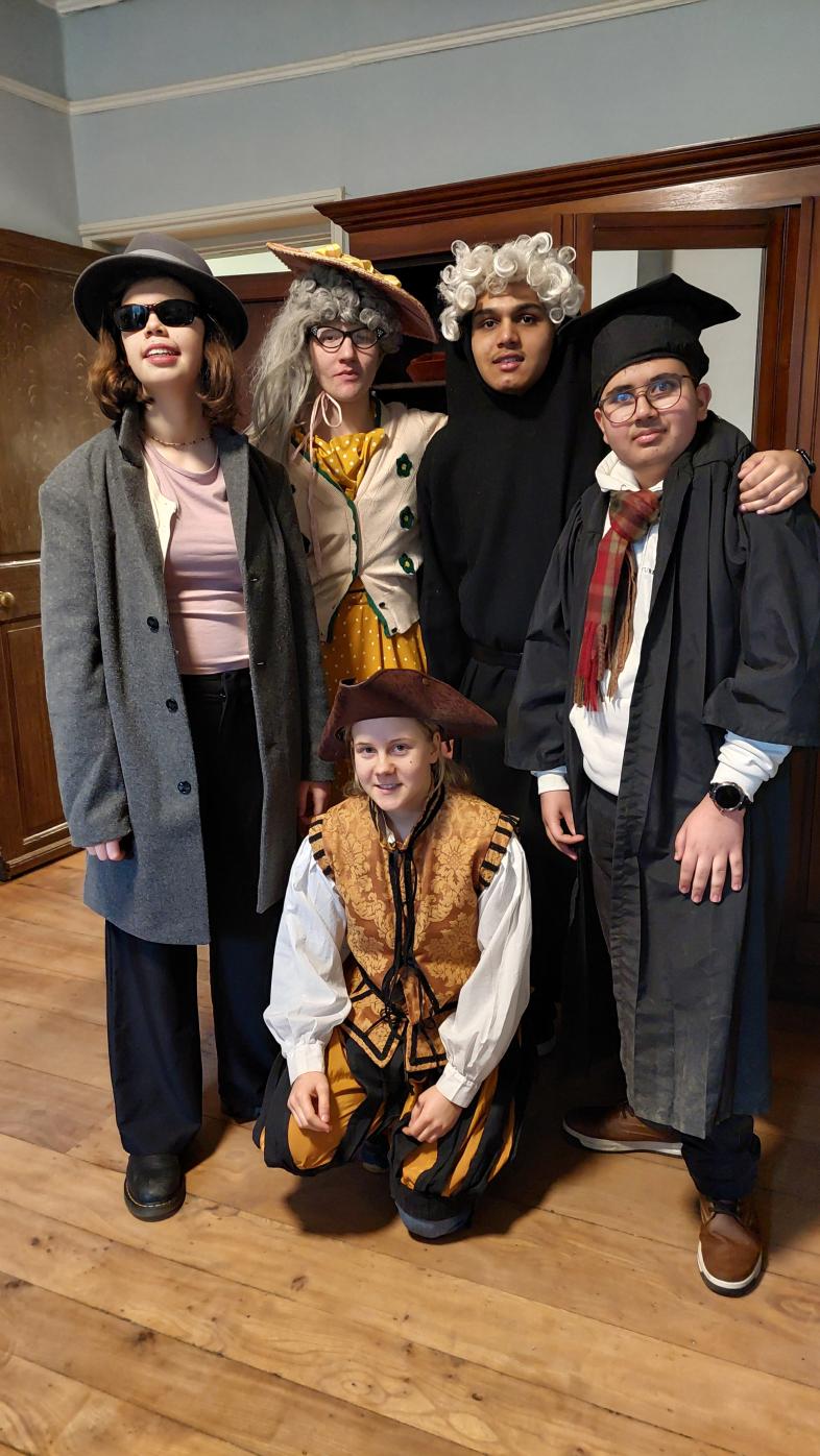 Fünf Schüler*innen in historischen Kostümen stehen und knien in einem Raum mit Holzboden vor einem Holzschrank.