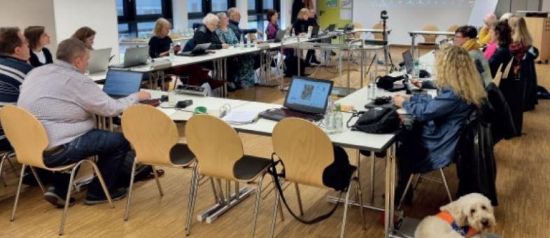 Rund 20 Teilnehmende der Tagung sitzen im Bielschowskyraum, vor ihnen Tische mit Laptops. Über einen Beamer sind auch die online zugeschalteten Menschen zu sehen.