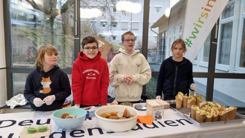 Die Sechstklässler Malte, Emilian, Max und Johannes stehen an ihrem Verkaufstisch in der blistaMensa und bieten Muffins und Wraps an.