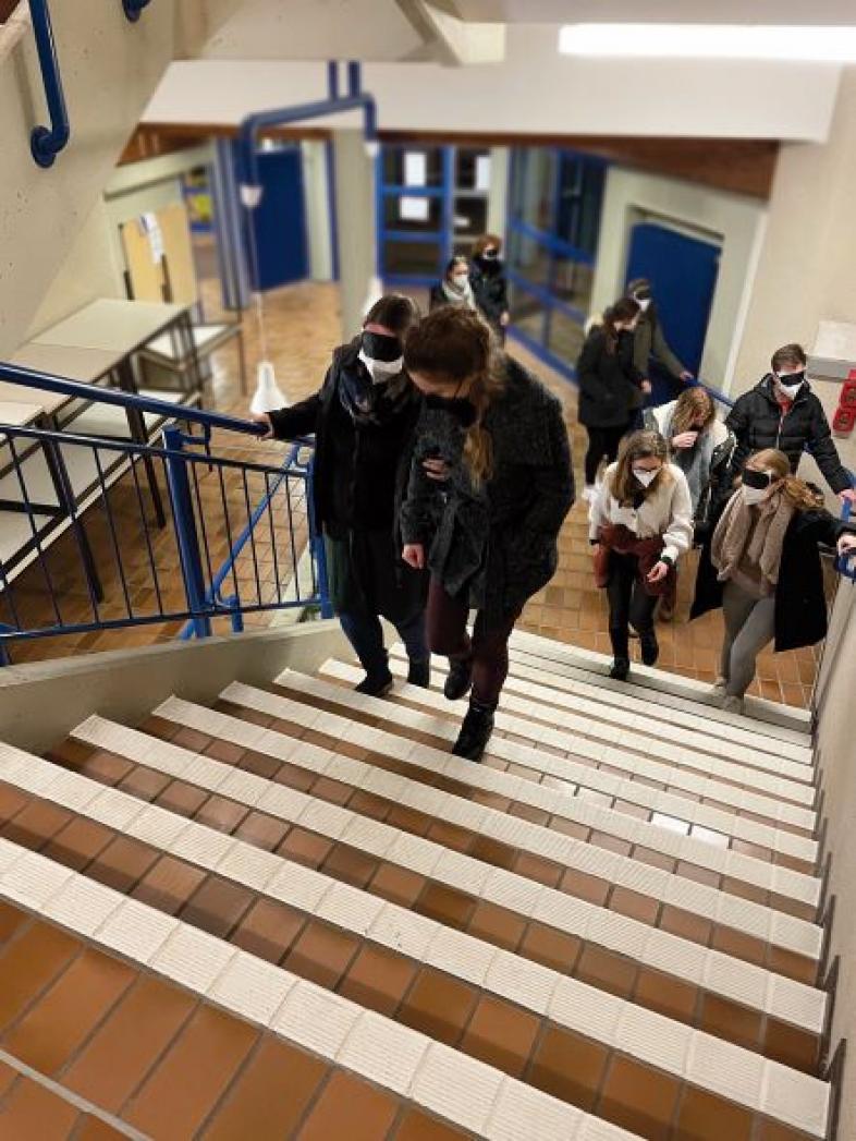 Studierende in Zweiergruppen gehen eine gut markierte Treppe hinauf. Eine Person hat die Augen verbunden, die zweite Person begleitet.