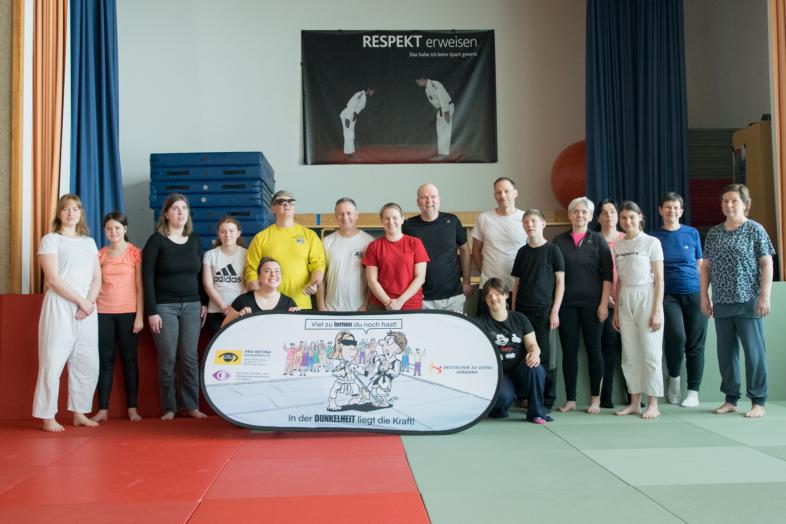 Die buntgemischte Gruppe aus 16 Menschen jeden Alters steht in der Gymnastikhalle der blista vor einem Banner von Pro Retina, DBSV und Ju-Jutsu-Verband. Darauf steht: „In der Dunkelheit liegt die Kraft.“