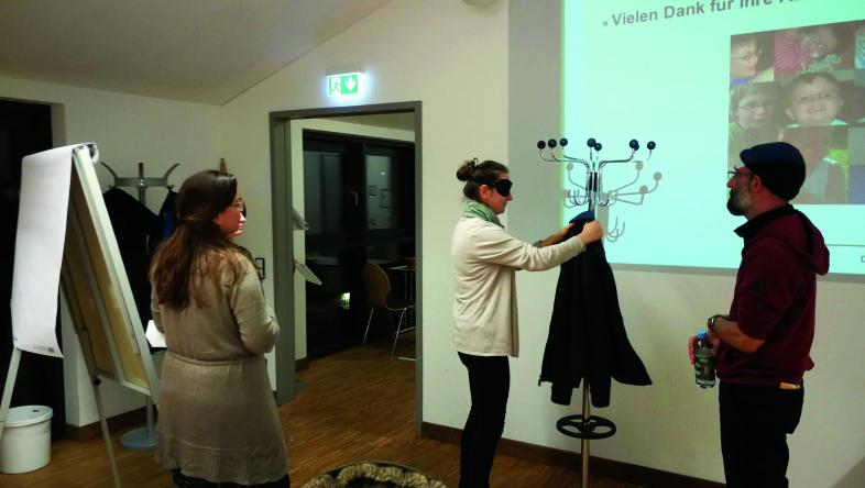 Eine Teilnehmerin mit Augenbinde nimmt eine Jacke von einem Garderobenständer. Rechts neben ihr steht der Teilnehmer, der ihr verbale Anleitungen gibt.