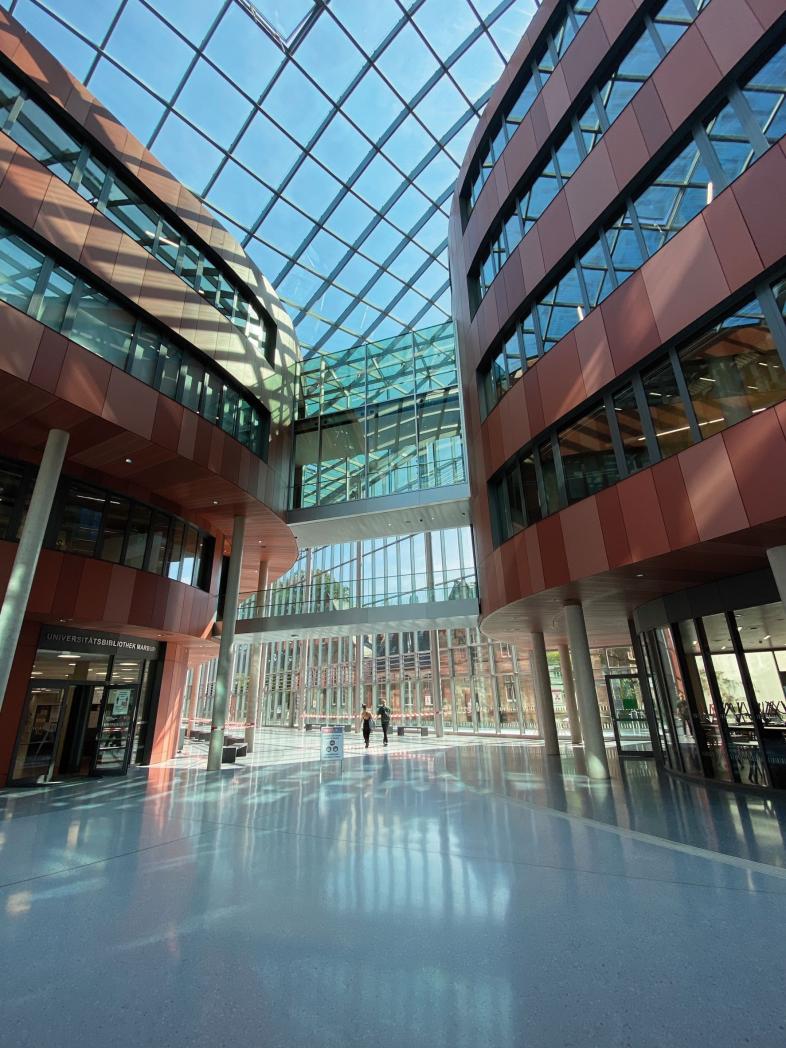 Das riesige Atrium der neuen Uni-Bibliothek der Philipps-Universität Marburg hat einen spiegelenden Boden und erstreckt sich über die gesamte Höhe des mehrgeschossigen Baus. Die Seitenwände bestehen aus Glas und rötlichem Beton, der Boden ist hellgrau.