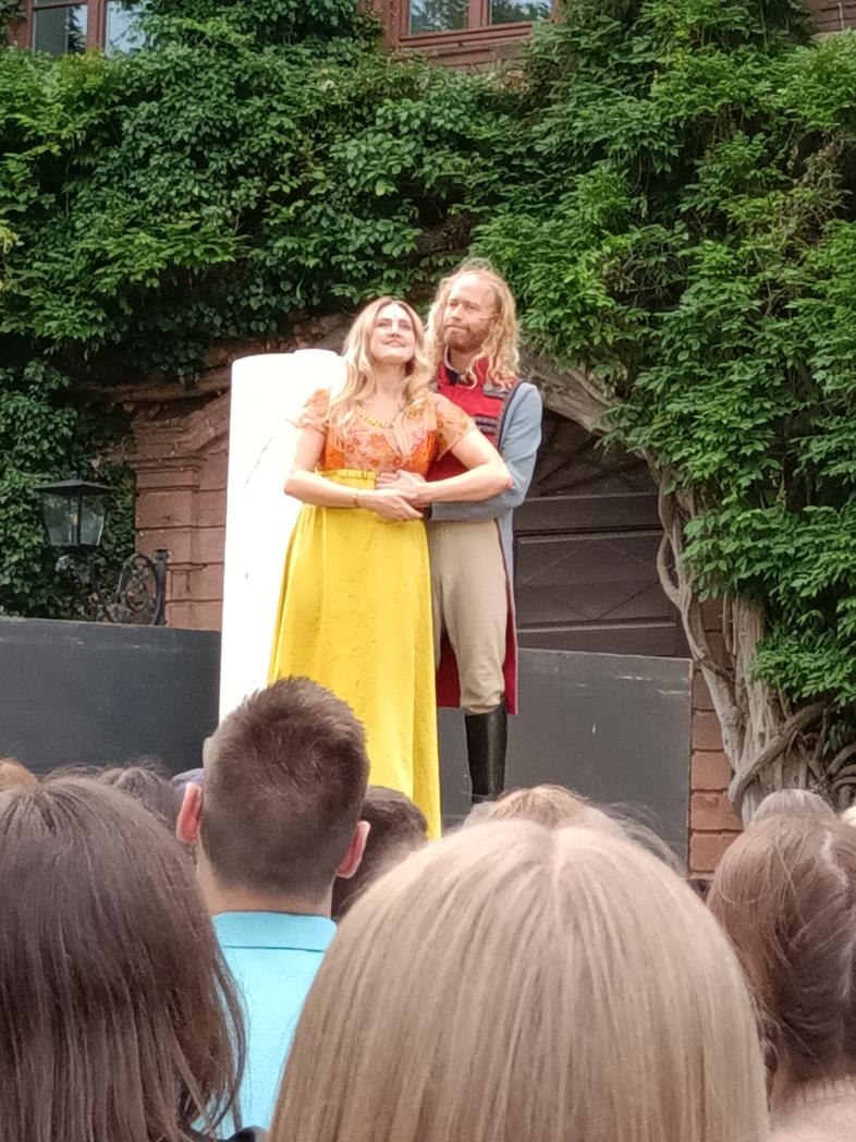 Ein Schauspieler steht hinter einer Schauspielerin auf einer Freilichtbühne, er hat seine Hand auf ihrer Taille, sie schauen Richtung Publikum.