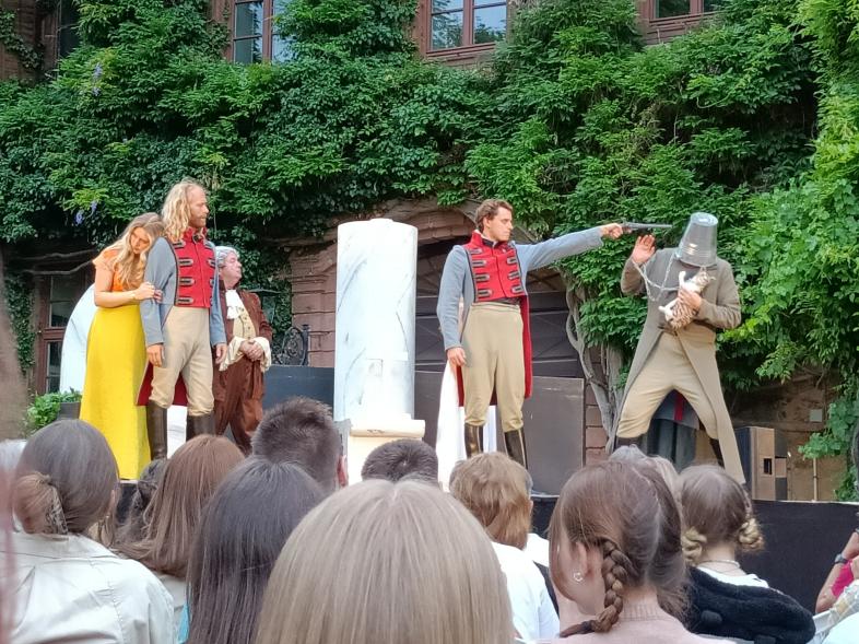 5 historisch gekleidete Schauspieler*innen. Einer der Schauspieler hält einem anderen eine Waffe an den Kopf, welcher wiederum einen Eimer auf dem Kopf hat und eine Katze im Arm trägt.
