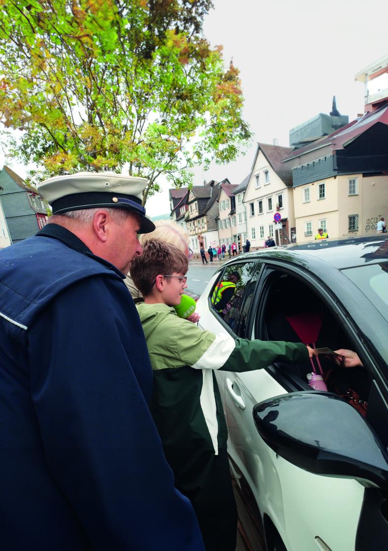 Gemeinsam mit einem Polizisten überreicht ein blista-Schüler einer Person in einem Auto eine grüne Karte für rücksichtsvolles Fahrverhalten. 
