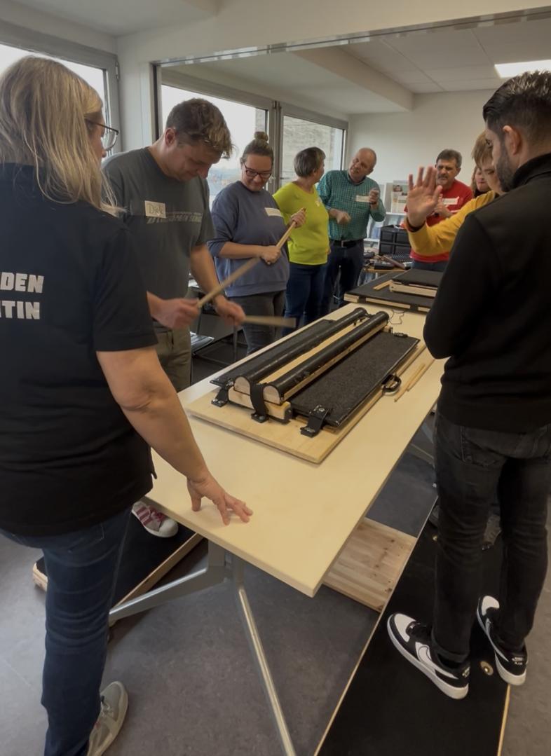 In den Räumen der Beratungsstelle musizieren mehrere Teilnehmende auf den Elektro-Tischtrommeln, unterstützt von Assistenz.