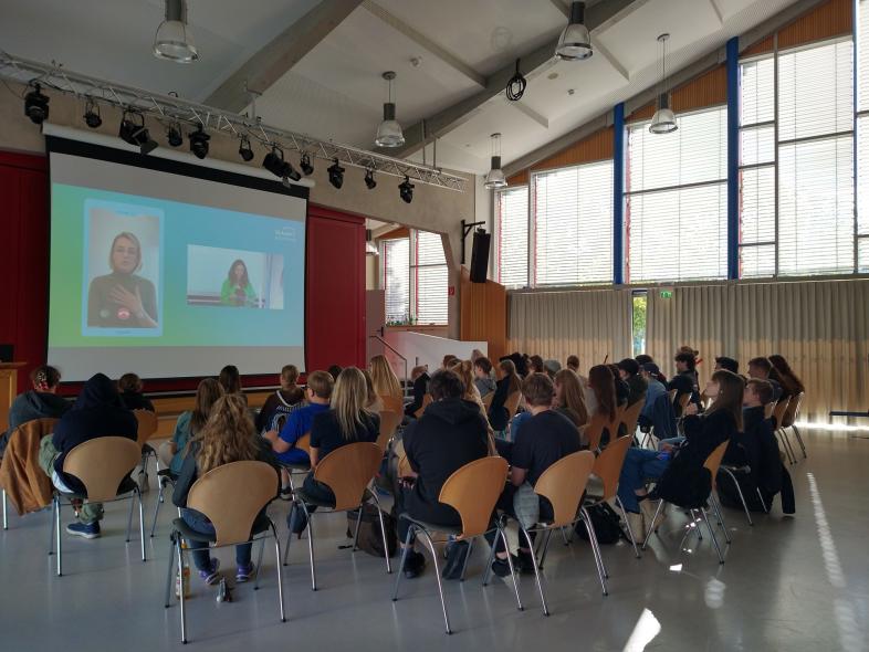 In der abgedunkelten Aula sitzen Schüler*innen auf Stühlen vor einer großen Leinwand und folgen einer Online-Veranstaltung.