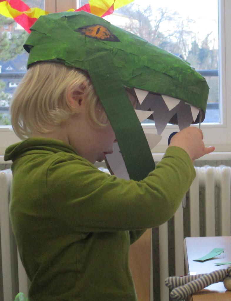 Ein Kindergartenkind trägt einen großen Drachenkopf aus Pappmaché auf dem Kopf und scheint durch das offene Maul des Drachen konzentriert zu arbeiten.